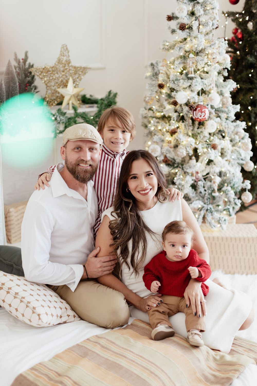 A family poses in front of a white Christmas tree decorated with ornaments and stars while sitting on a cozy bed.