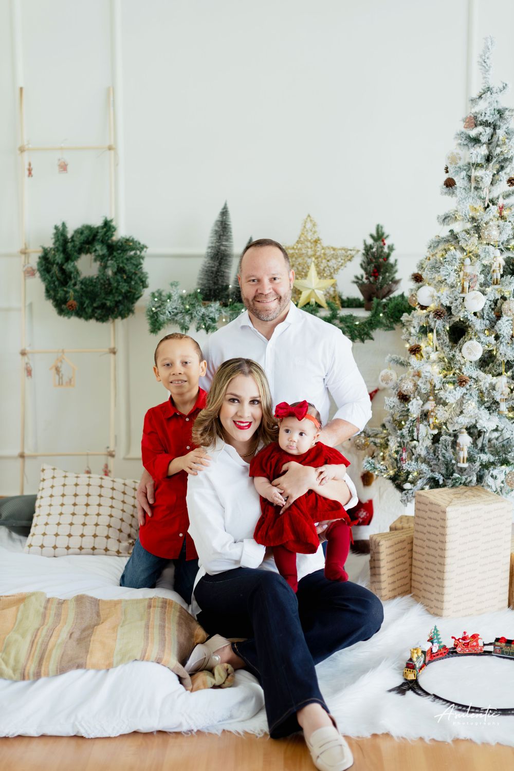 Family dressed in red and white poses together on couch with decorated Christmas tree in background.