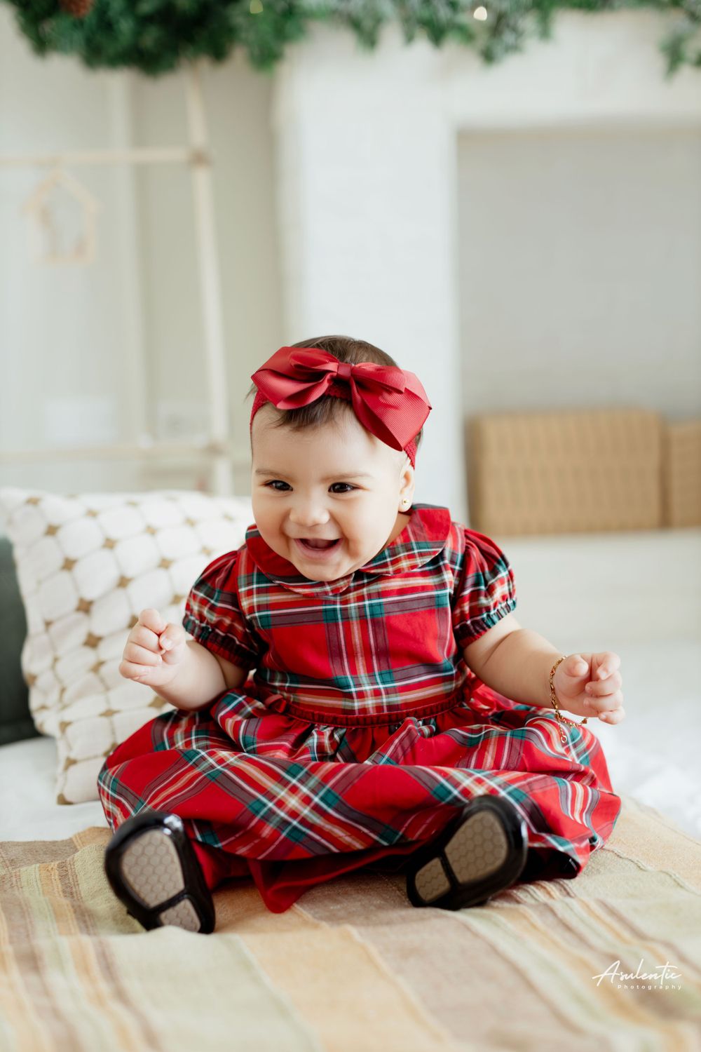 Baby in red plaid holiday dress with bow sitting on couch near Christmas decorations.