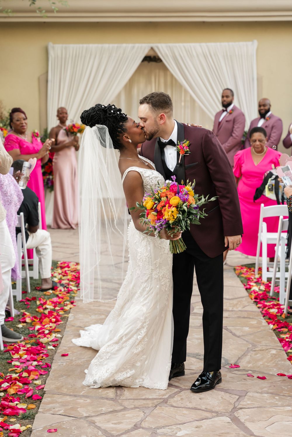 Newlyweds share a kiss at outdoor ceremony with pink rose petals scattered along aisle and guests in bright pink dresses.
