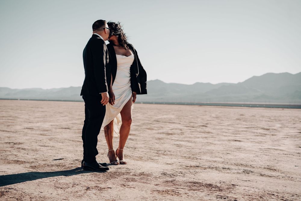 A romantic couple in formal attire stands together on a scenic desert salt flat at sunset.