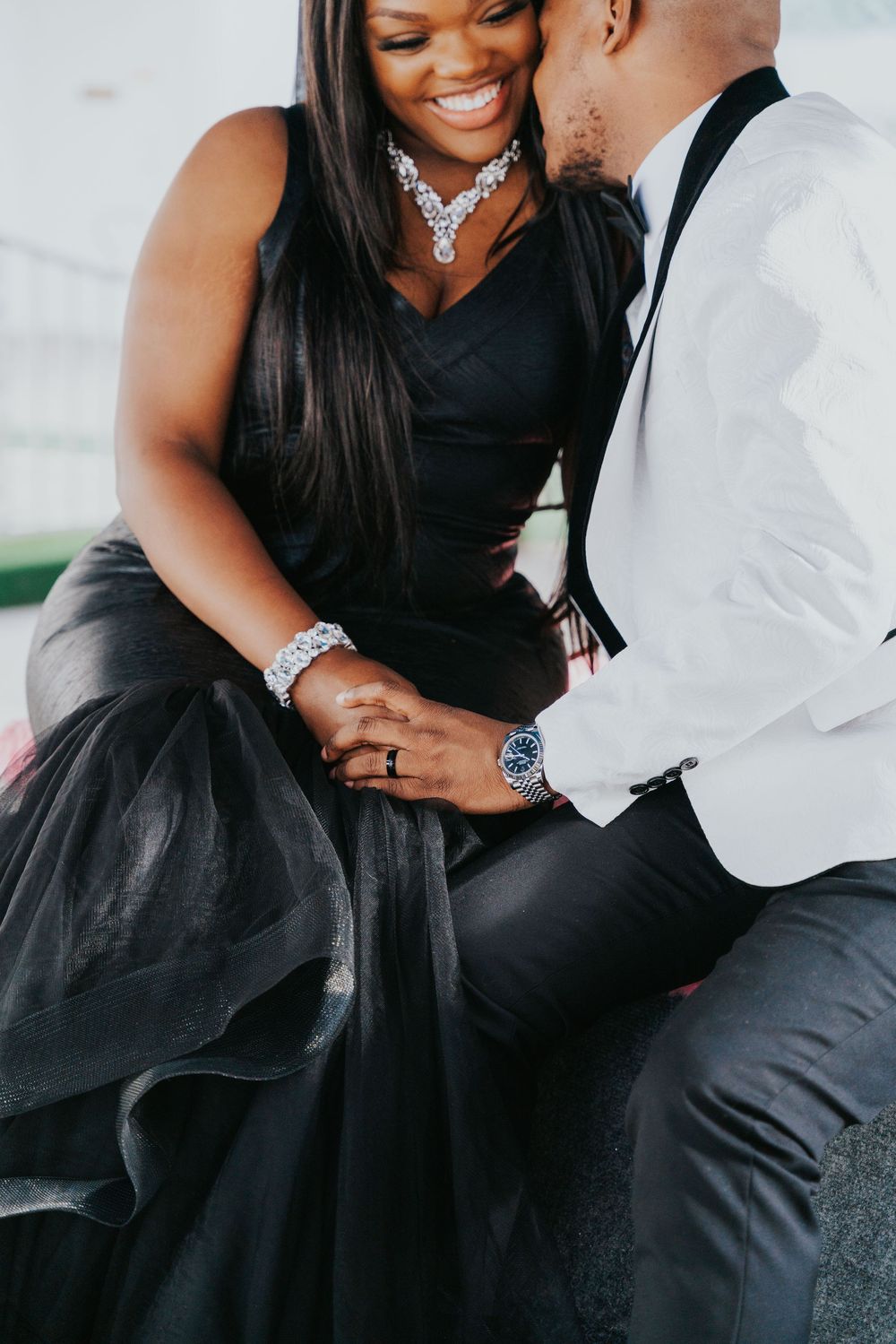 A couple in formal attire shares an intimate moment, with the woman wearing a black dress and elegant jewelry.