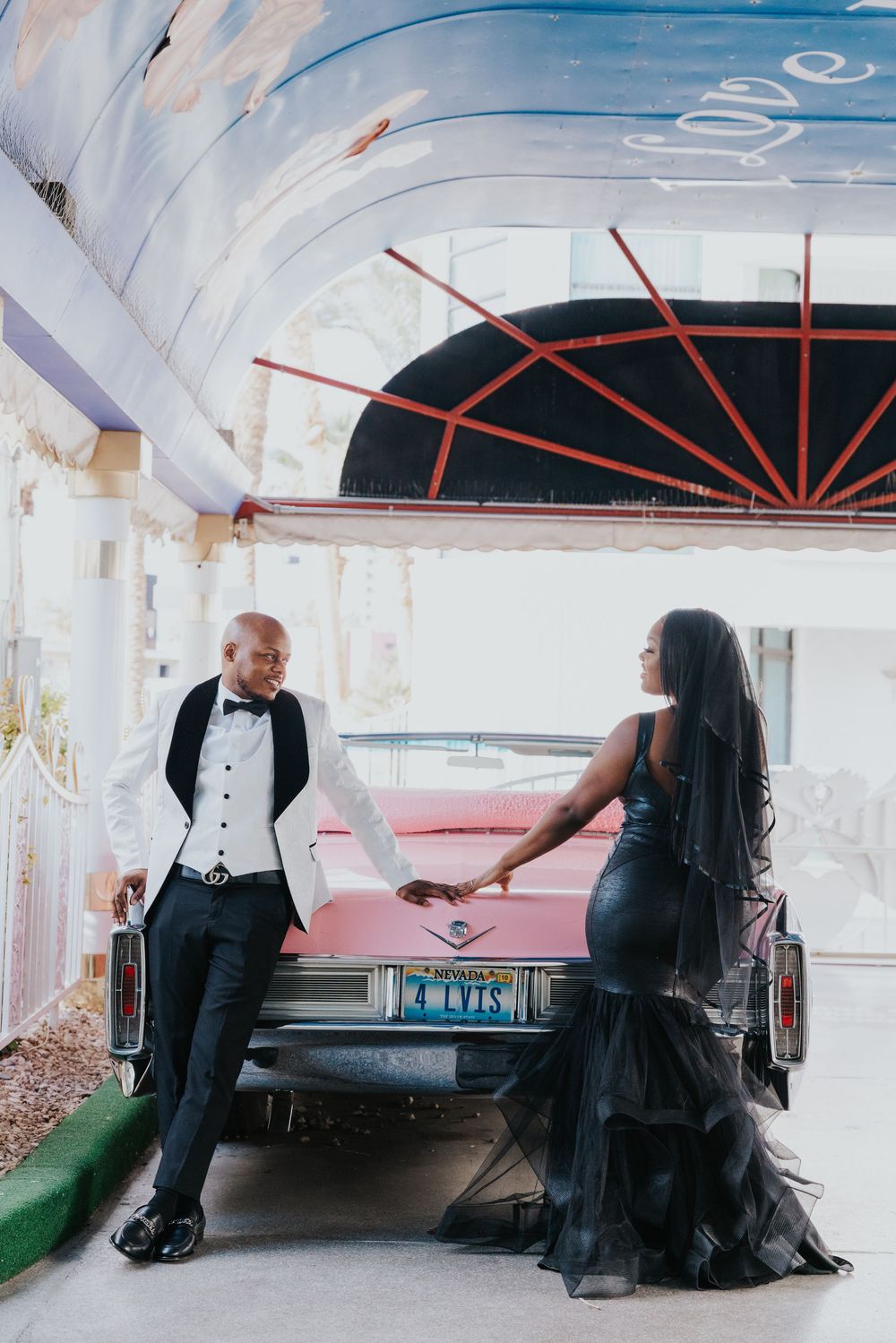 Elegant couple in formal black attire pose with vintage pink Cadillac at stylish venue. A little white wedding chapel elopement photography.