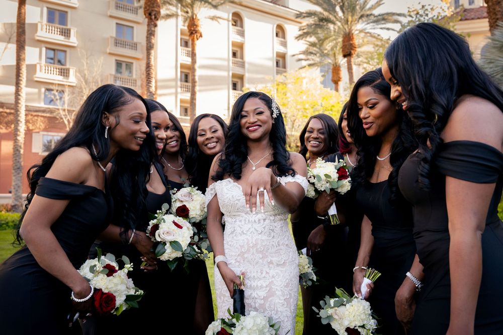 Wedding party members in black dresses gather around someone in white for joyful group photos at an outdoor venue.