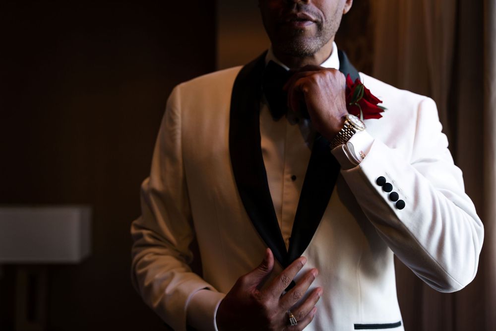 A person wearing a white tuxedo jacket with black lapels and red boutonniere adjusts their bowtie in dim lighting.