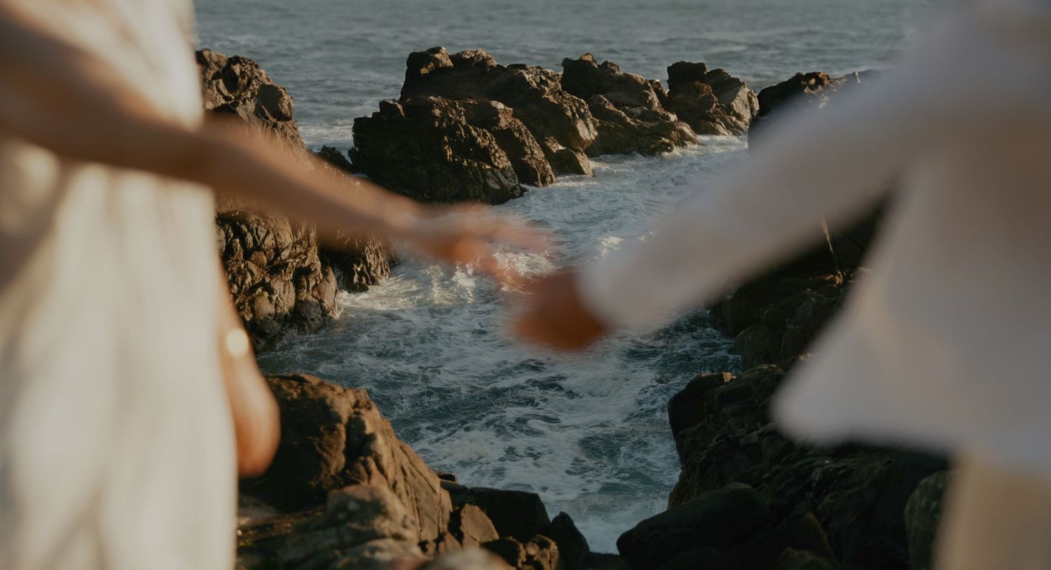 Hands clasped together against a scenic rocky coastline with crashing ocean waves at sunset.