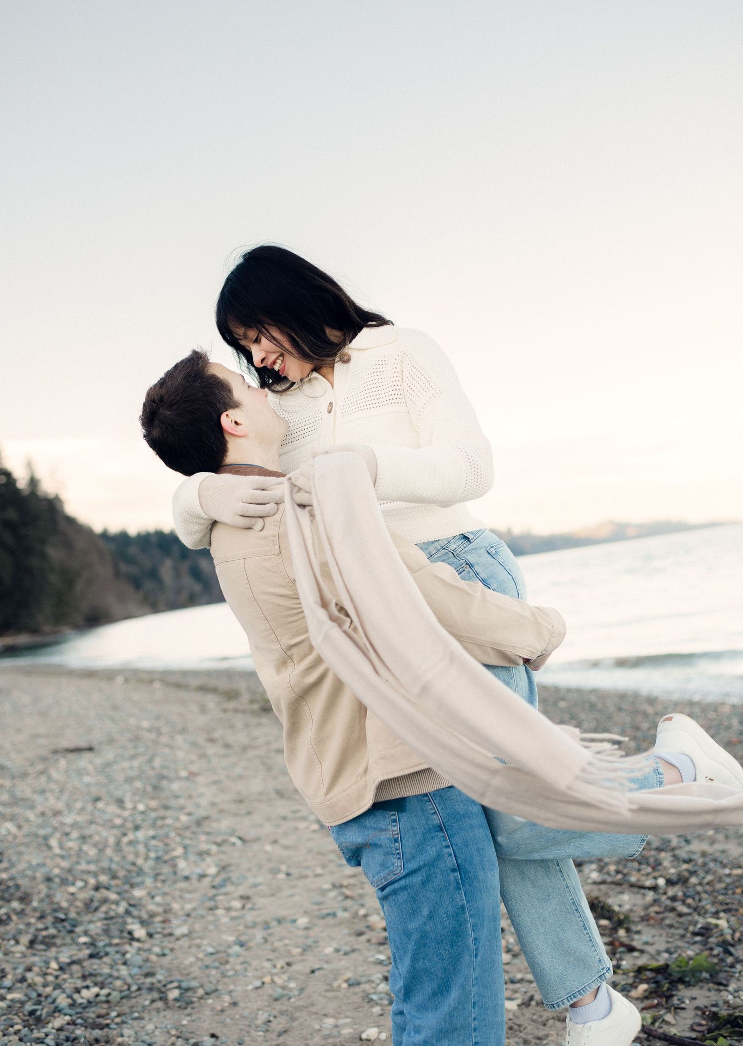 A couple embraces on a rocky beach shore during sunset, wearing casual denim and light colored sweaters.
