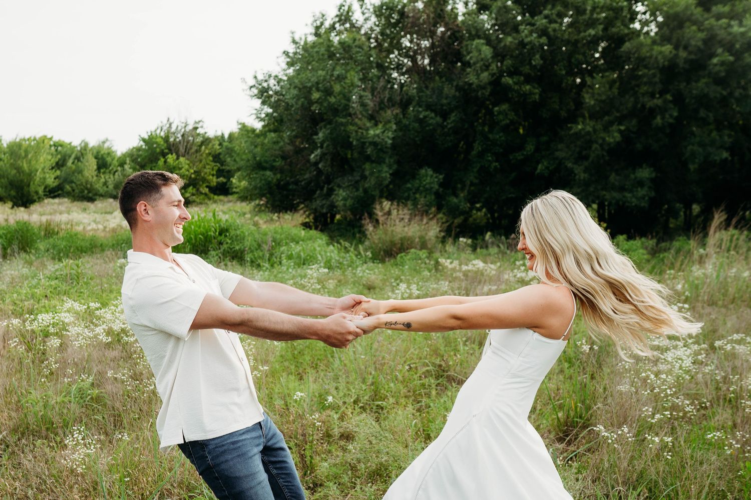 A couple in white outfits holds hands and spins playfully in a sunny meadow with trees in the background.