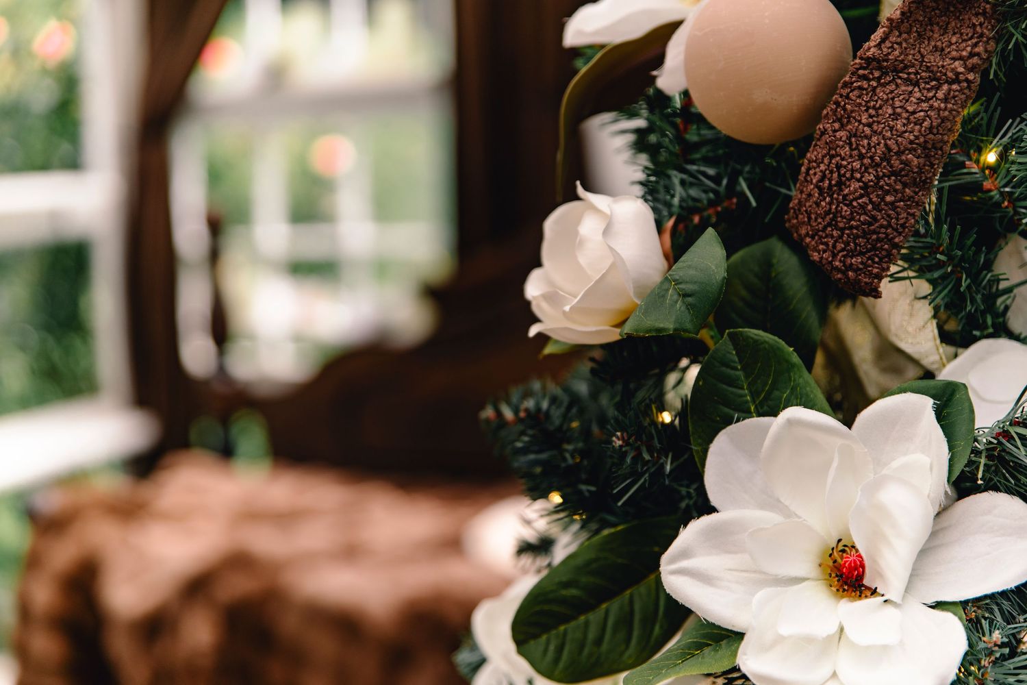 White magnolia blossoms and green leaves on a festive Christmas garland with twinkling lights, with a cozy blurred window scene behind.