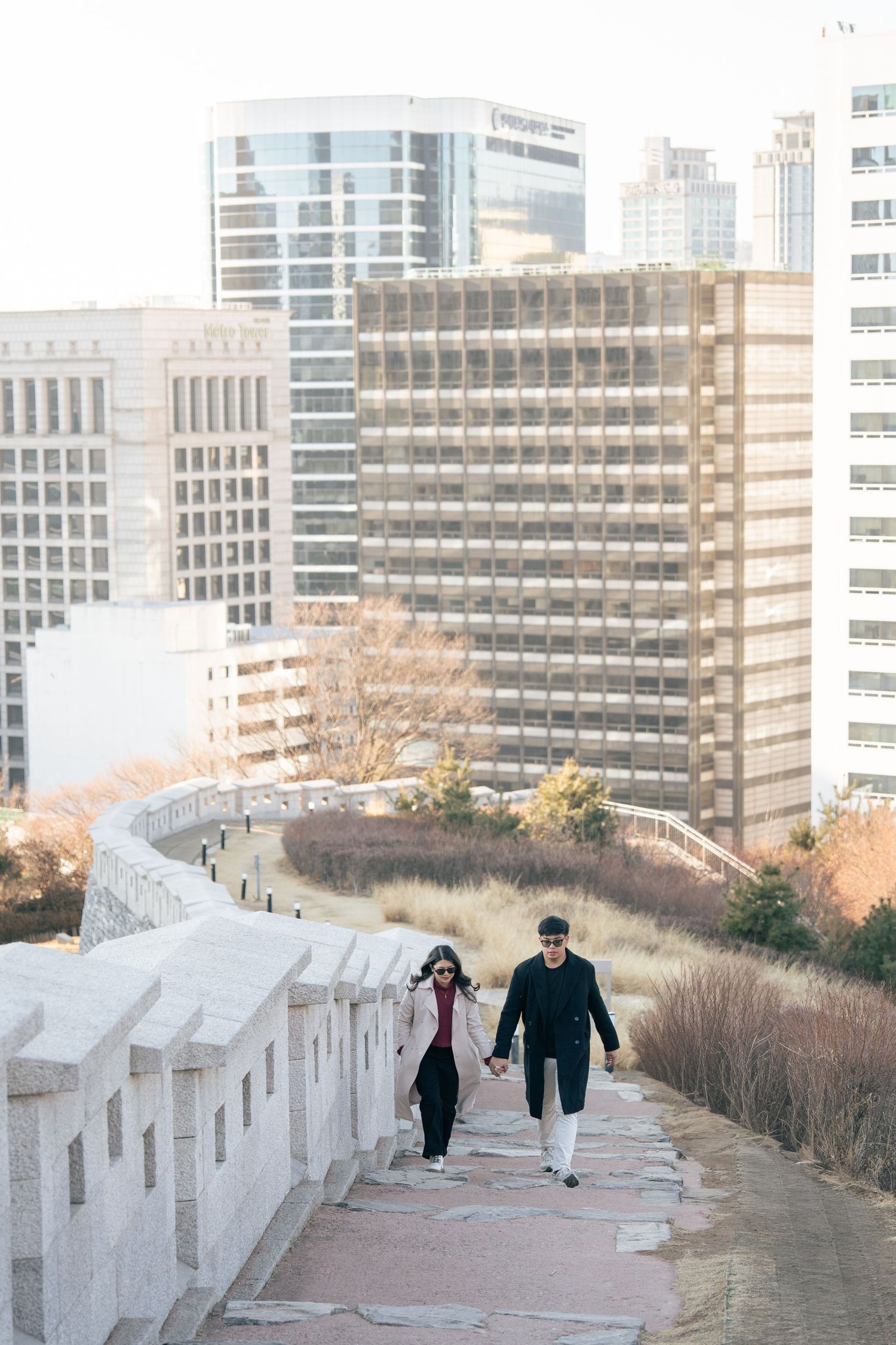People walking along a white stone wall pathway with urban buildings in the background on a winter day.