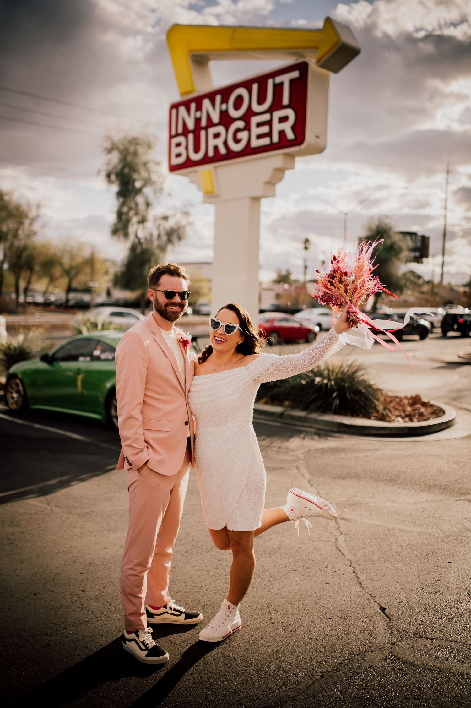 A couple in matching pink outfits poses playfully outside an In-N-Out Burger restaurant on a sunny day.