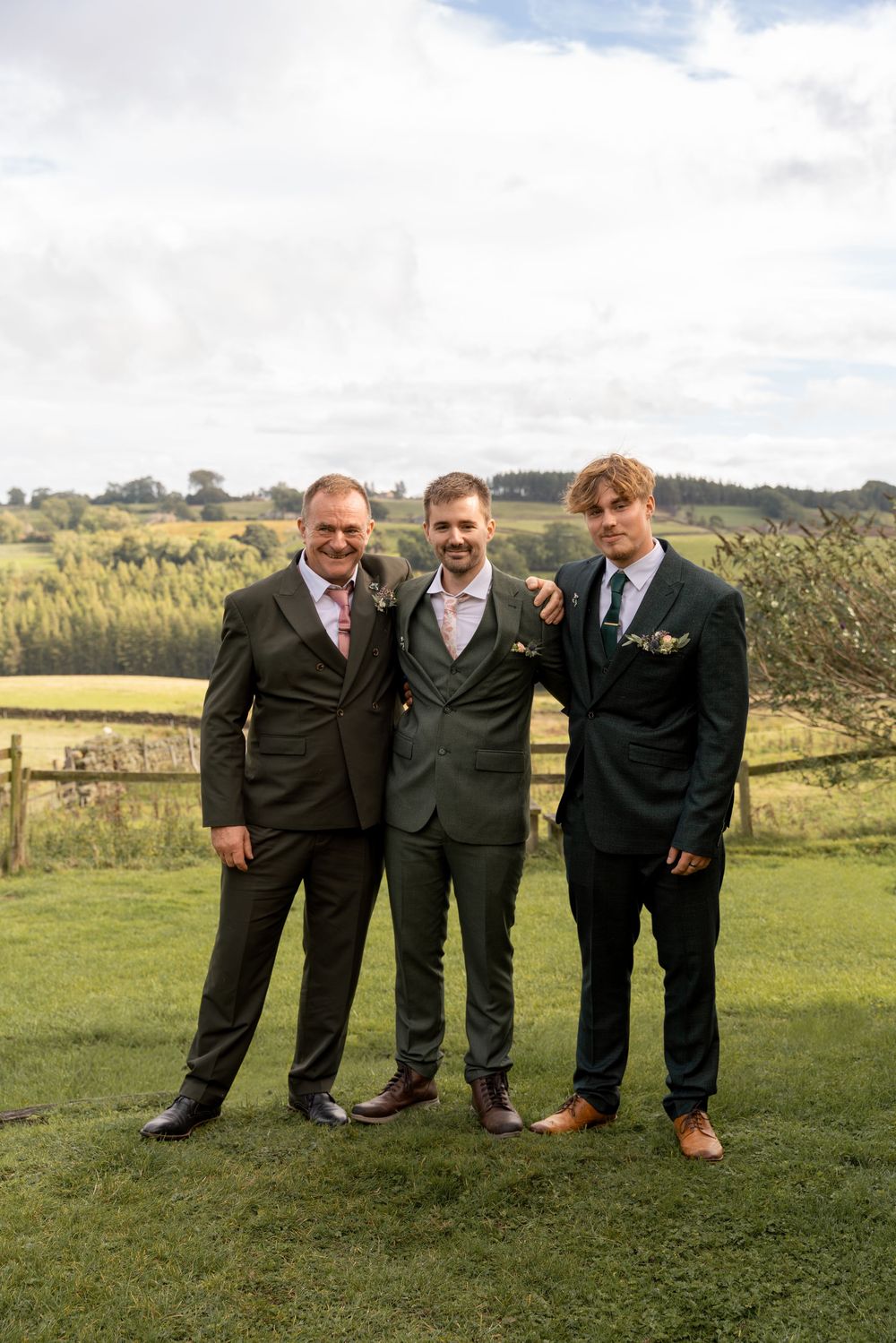 Three groomsmen in dark suits pose together on a grassy hillside with scenic countryside views in the background.