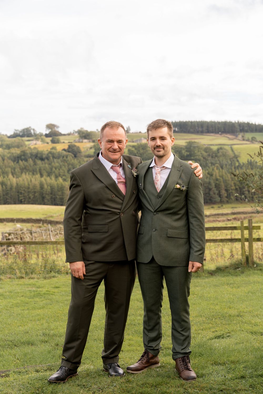 Two people in dark suits stand together on green grass with rolling hills and trees in the background.