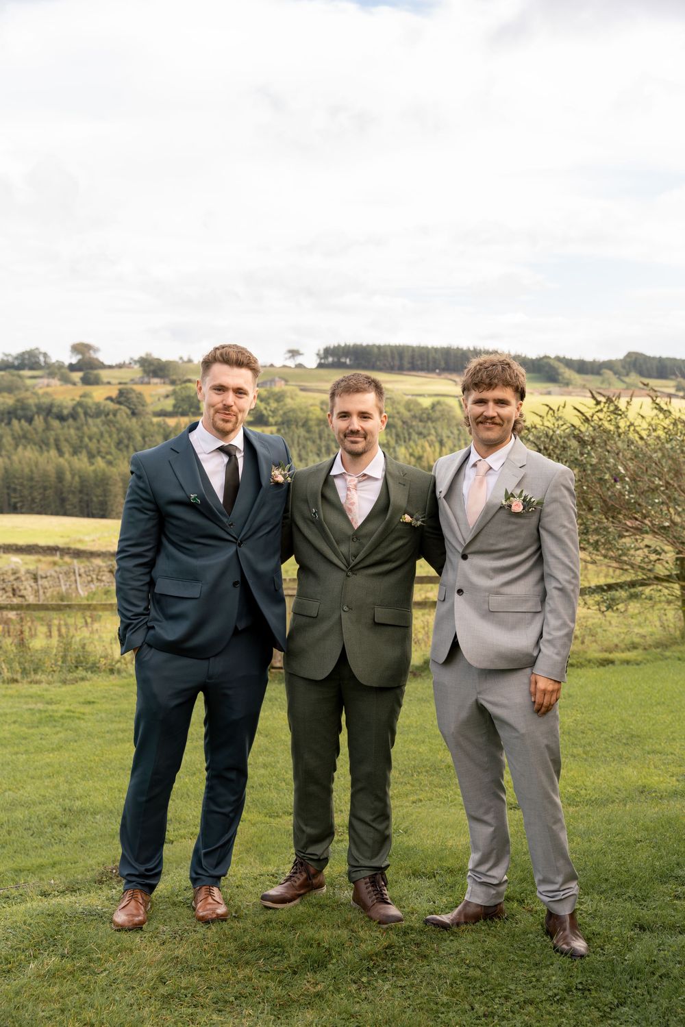 Three men in suits standing together on grassy hill with scenic countryside landscape in background.