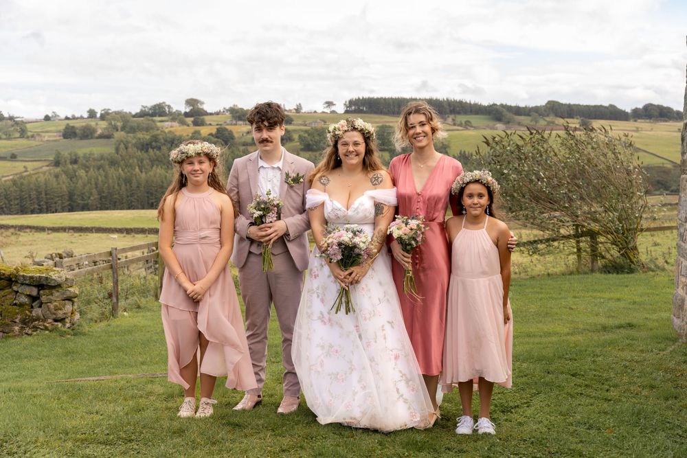 A group of people in pink and blush-colored formal attire pose together on a grassy hilltop during a wedding celebration.