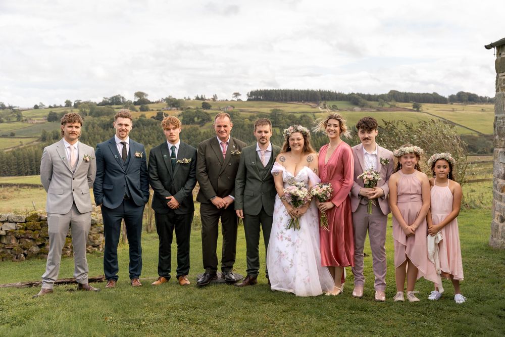 A wedding party stands together outdoors in formal attire against a scenic natural landscape backdrop.
