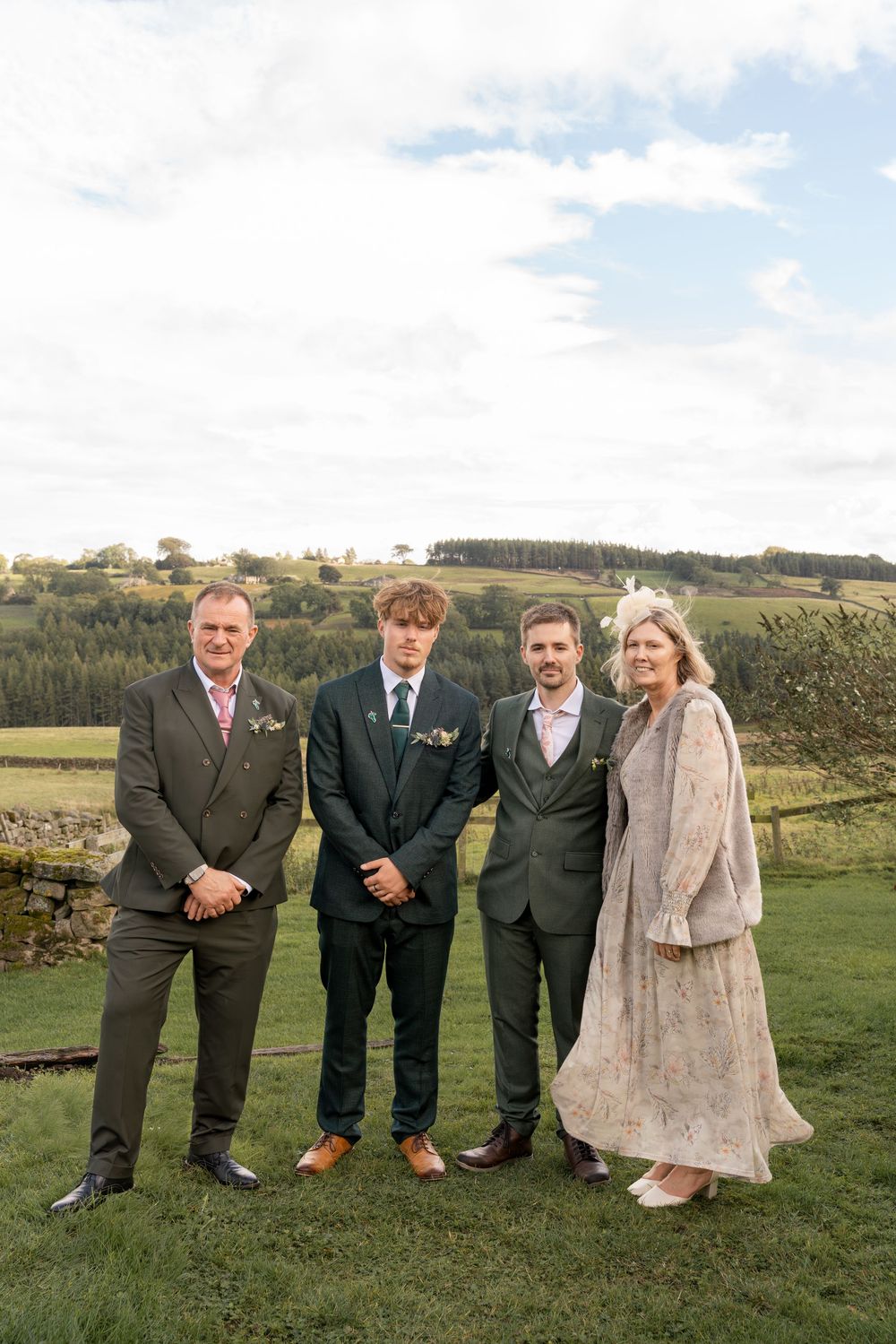 Wedding party members stand together on a grassy hillside in formal attire against a scenic countryside backdrop.