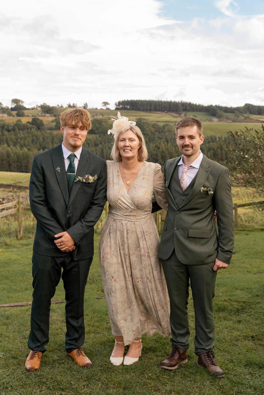 Three people in formal attire pose together on a grassy hillside with trees and rolling countryside in the background.