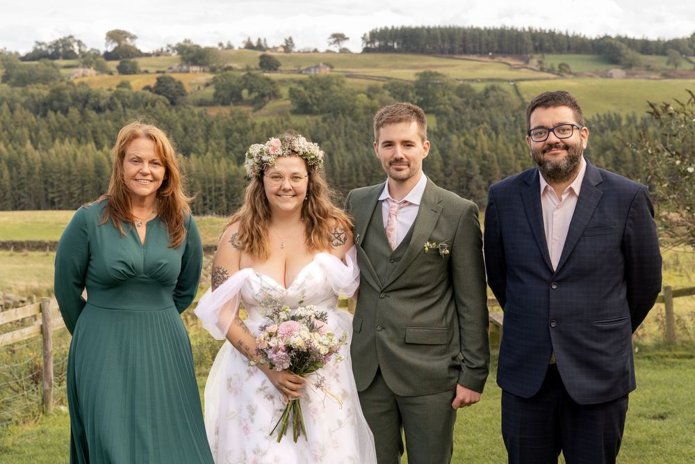 Wedding group photo with people wearing emerald green and formal attire posing outdoors in a rural setting.