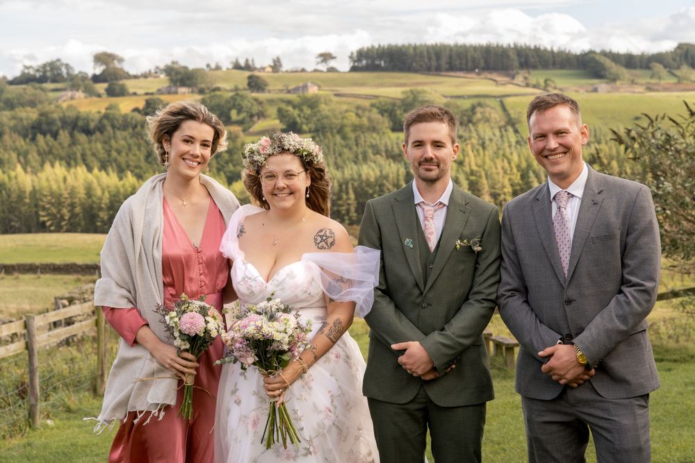 Wedding party poses together in outdoor countryside setting with rolling hills and natural landscape in background.