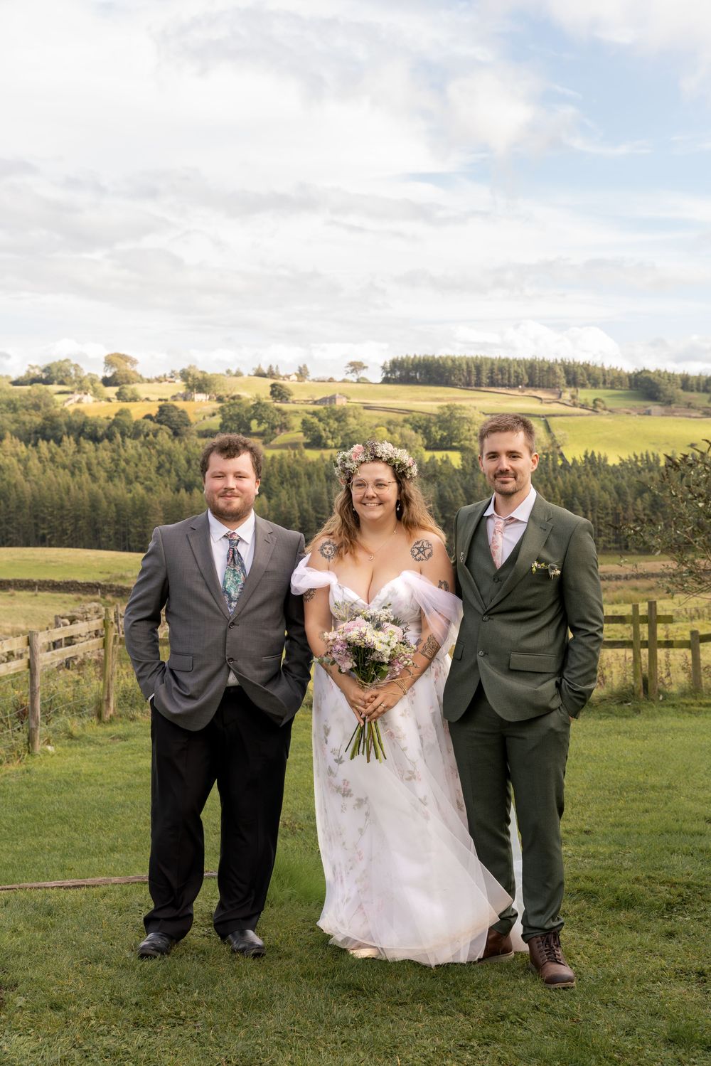 Wedding party poses in front of scenic rolling hills and countryside on a grassy field with wooden fencing.