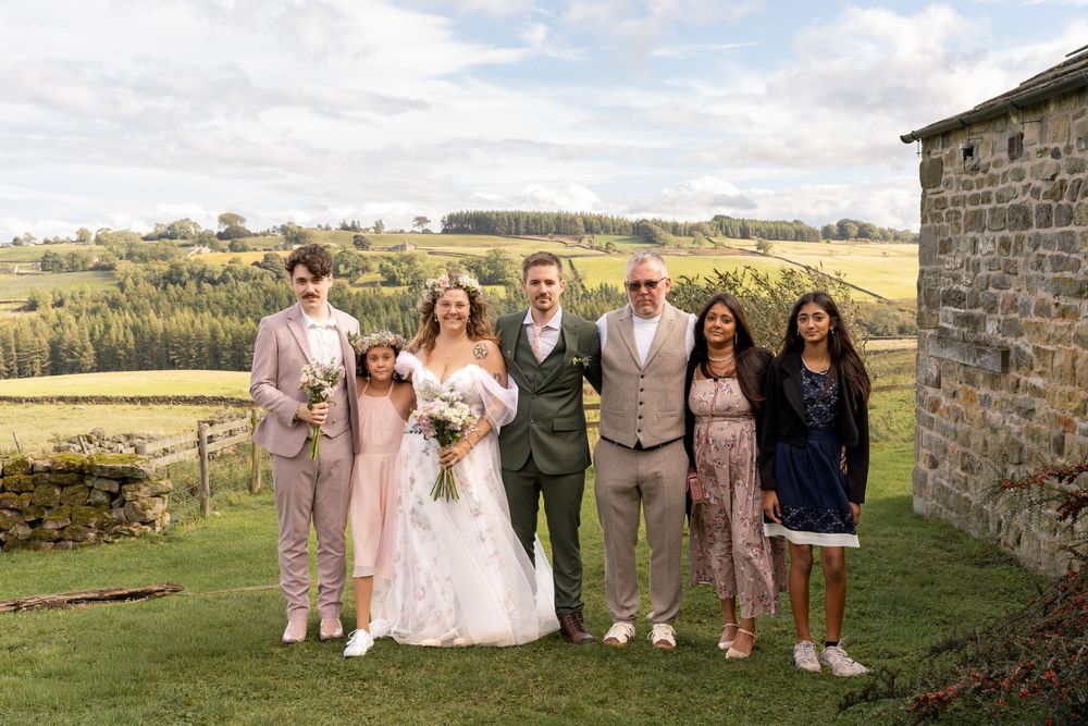 Wedding party poses together in front of a rustic stone wall with rolling countryside hills in the background.