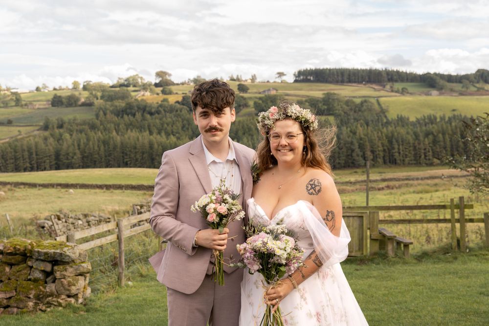 A newlywed couple poses in a scenic rural landscape with rolling hills and green pastures in the background.