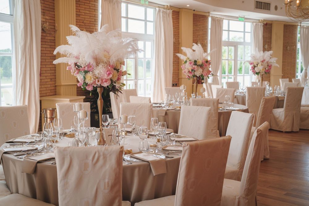 Elegant white wedding reception venue with round tables, white linens, and tall floral centerpieces in a bright sunlit room.