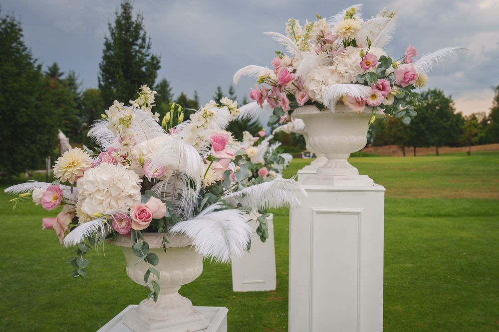Elegant white pedestals with ornate floral arrangements featuring pink roses and white blooms displayed on a grassy lawn.