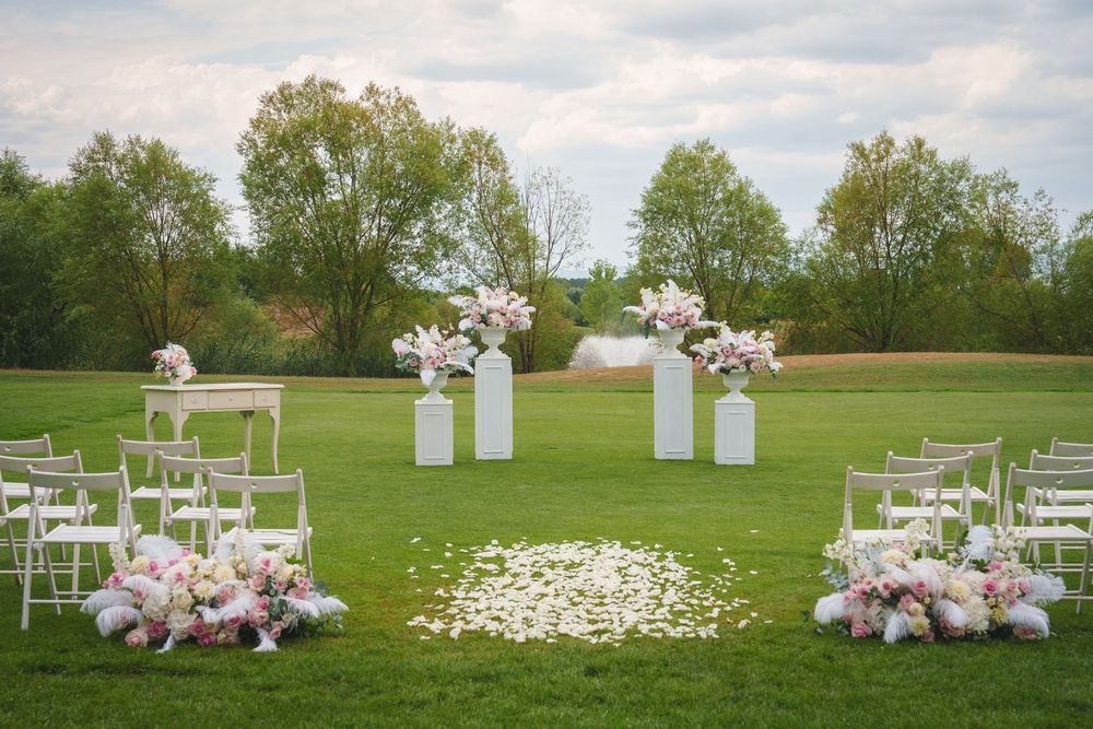 White wedding ceremony setup with chairs and floral decorations in a lush green field surrounded by trees.