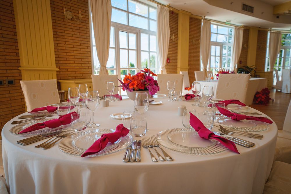 Elegant banquet hall with round tables set with white linens and pink napkins beside large windows with natural light.