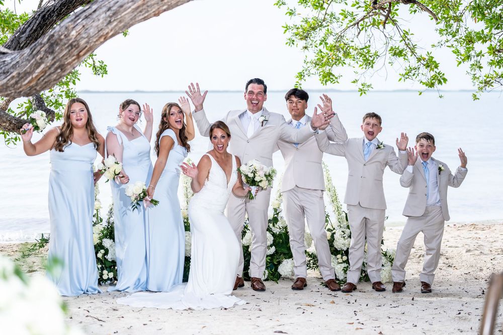 A wedding party in light blue and white attire poses joyfully on a tropical beach under a tree.