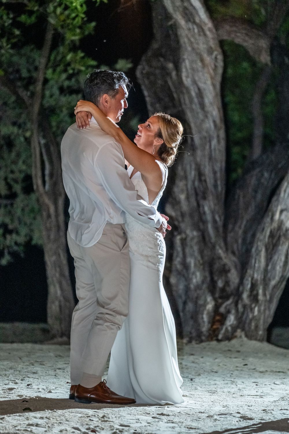 A couple in wedding attire sits together on a sandy beach surrounded by trees in a romantic moment.