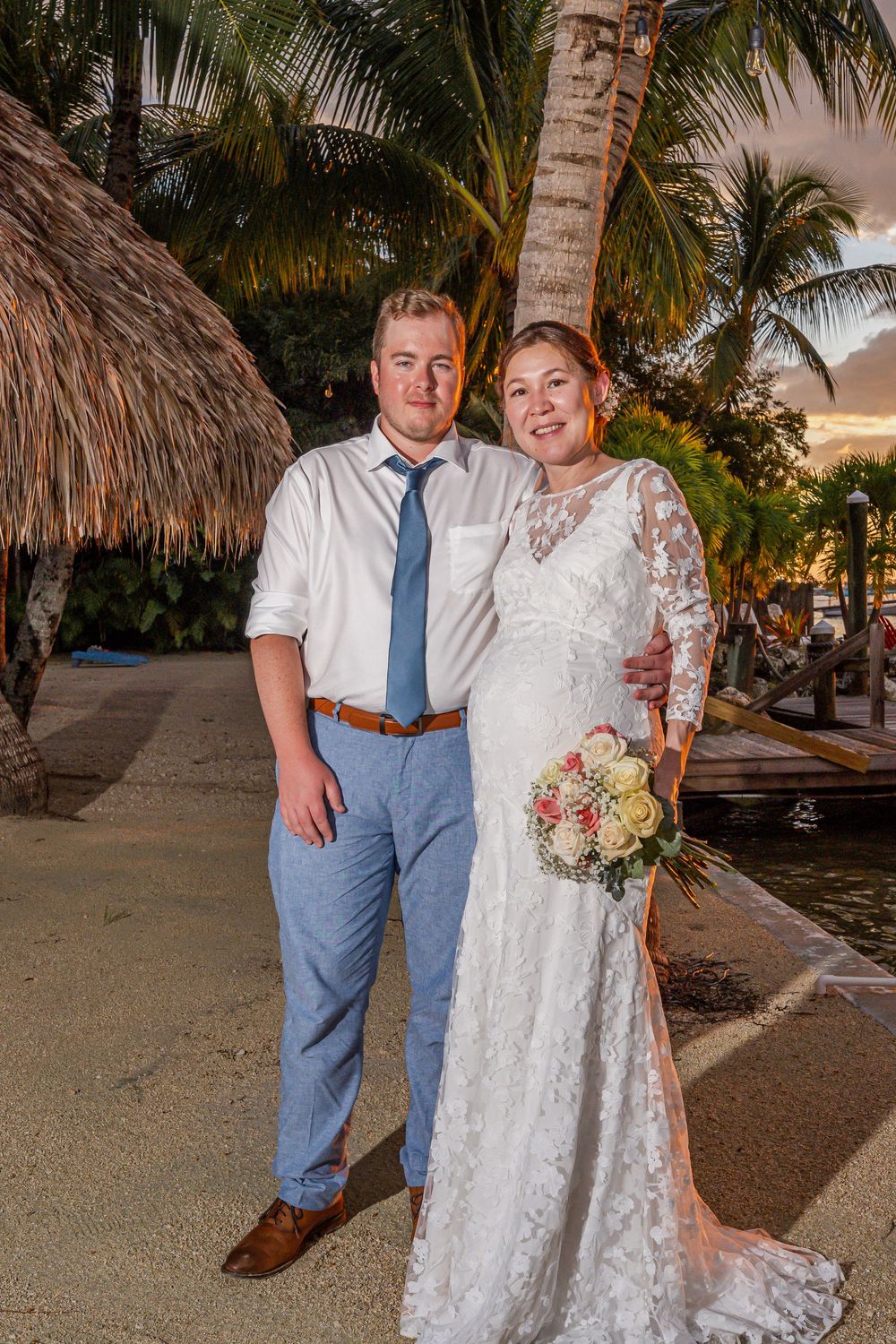 Newlyweds pose under palm trees at a tropical beach resort during sunset with thatched roof huts in background.