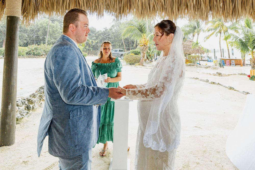 Wedding guests gather under a thatched-roof pavilion on a tropical beach for a celebration ceremony.
