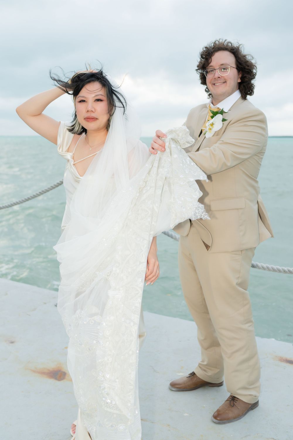 A couple in wedding attire poses playfully on a boat deck with the ocean and cloudy sky in the background.