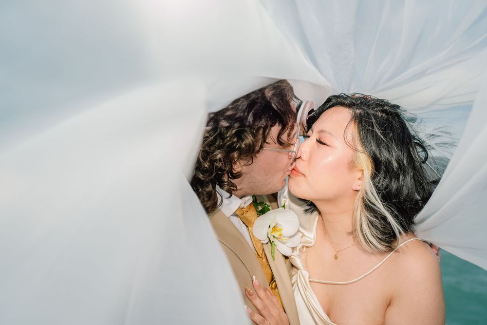 A romantic wedding kiss captured under a flowing veil with white flowers against a turquoise background.