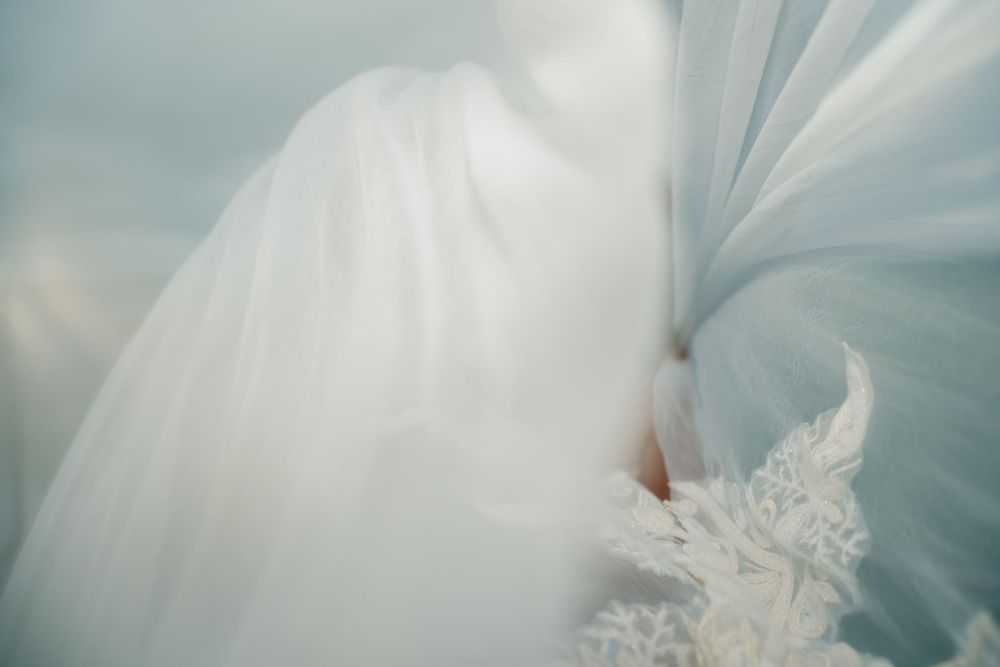 Close-up details of a white wedding dress showing delicate lace patterns and flowing fabric in soft light.