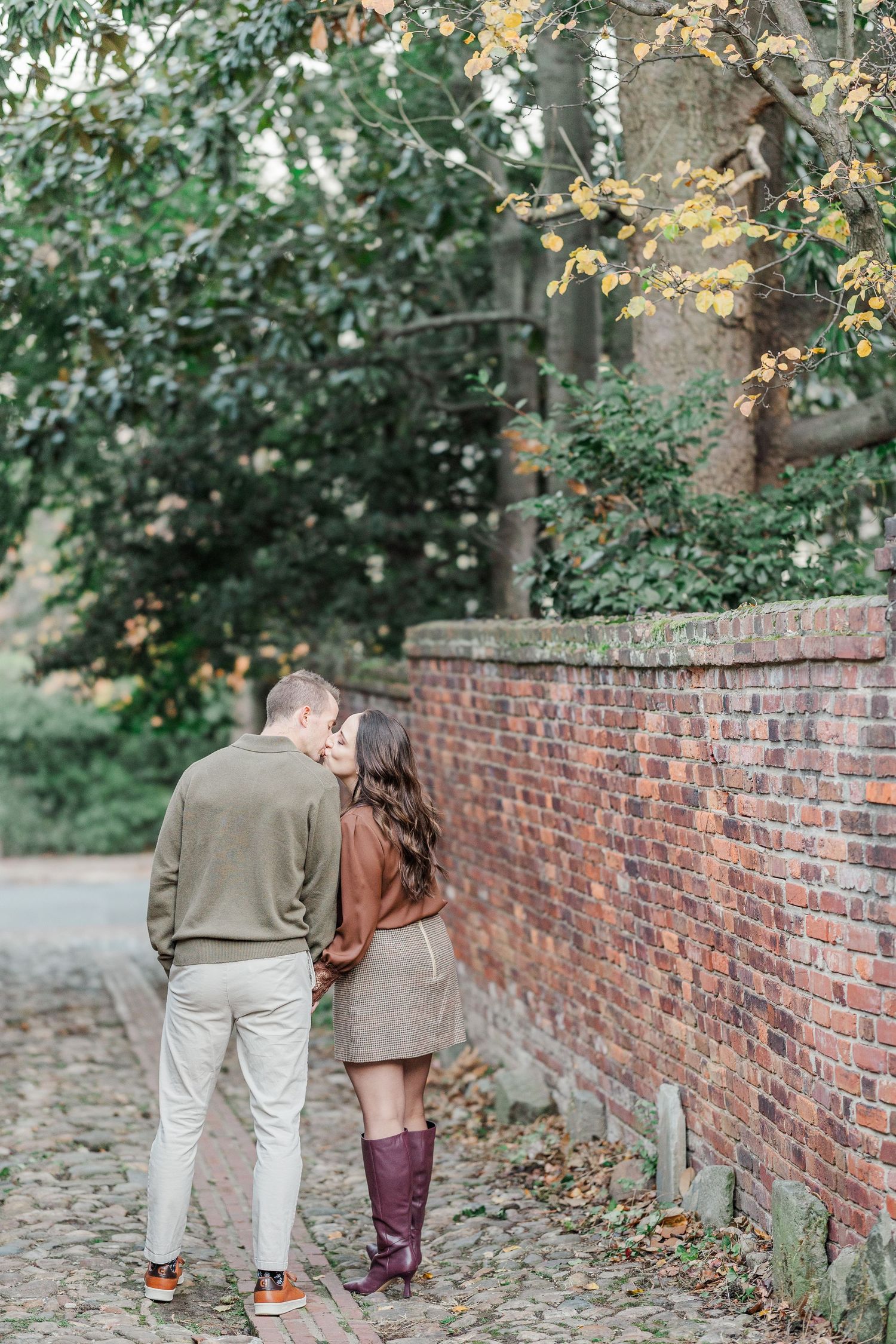 Two people walk together along a rustic brick wall path surrounded by lush green trees and foliage.