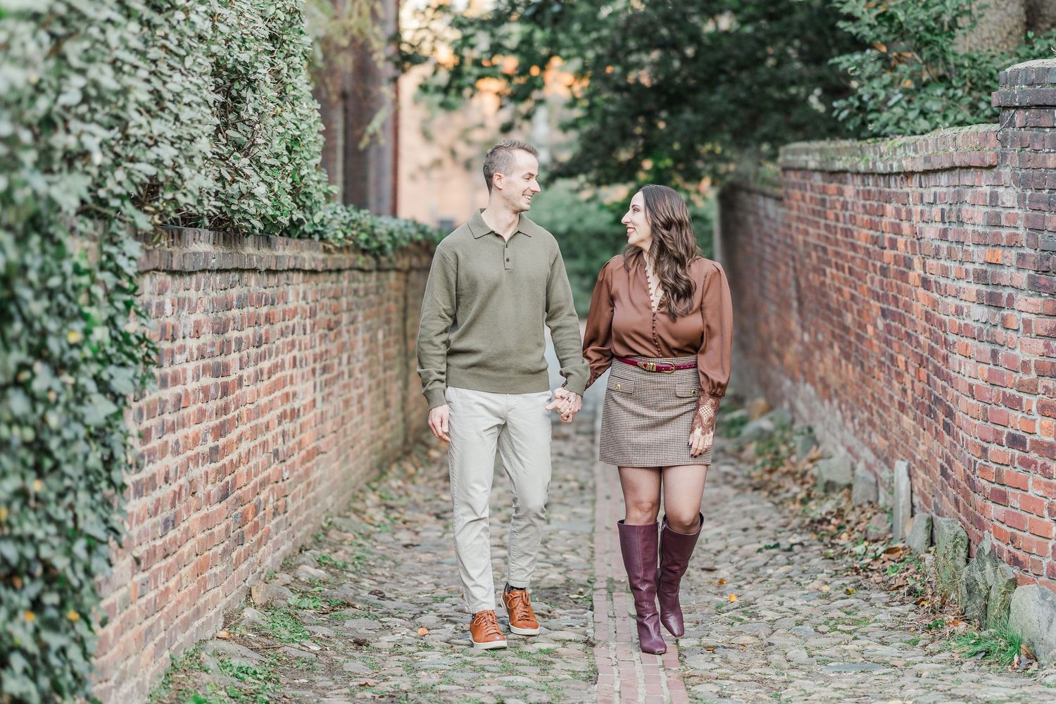 Two people walk along a narrow brick-walled garden path surrounded by green foliage.