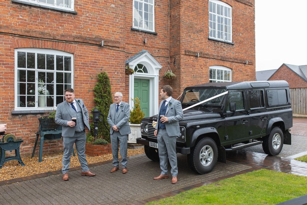 A group stands outside a traditional red brick house with a black Land Rover Defender parked in the driveway.