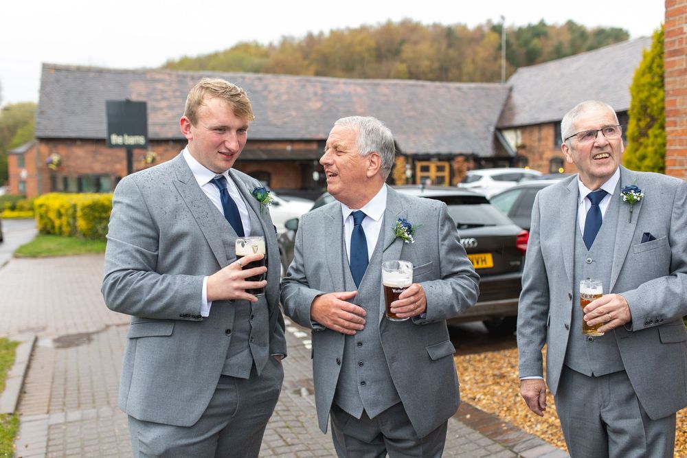 Three groomsmen in matching gray suits share drinks and conversation outside a rustic venue.