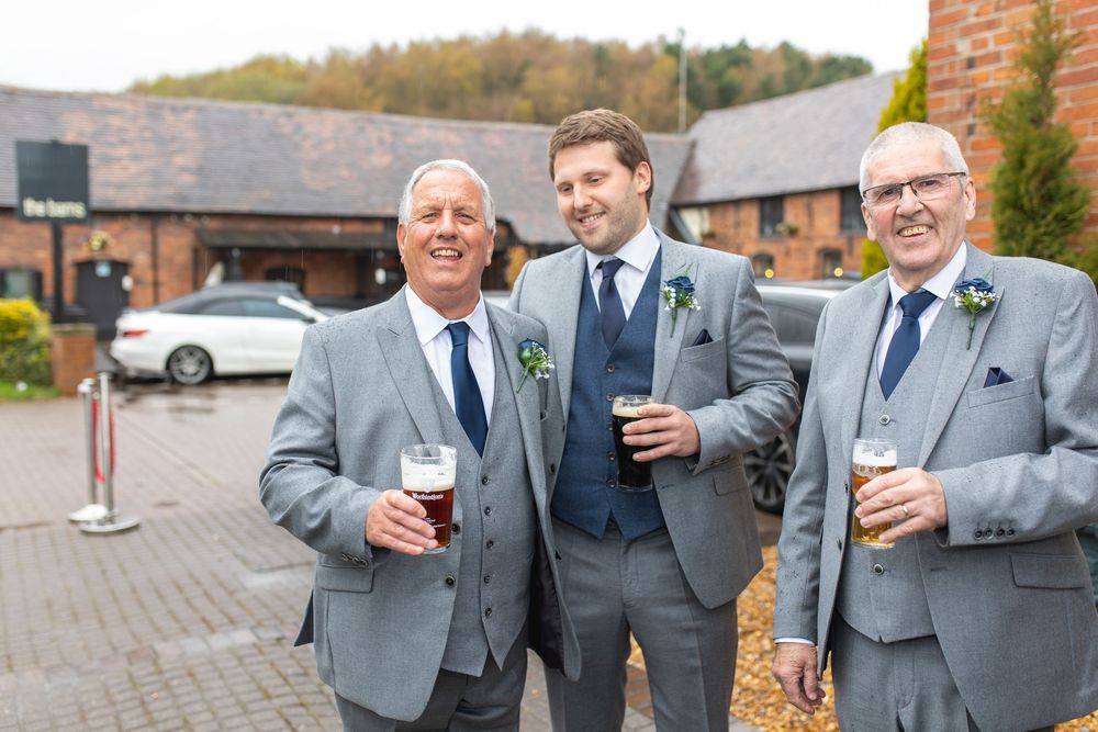 Wedding party members in matching gray suits toast with drinks during outdoor celebration.