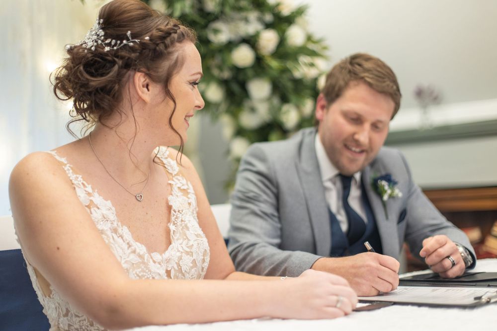 Wedding ceremony participants signing documents at a table with decorative floral arrangements.