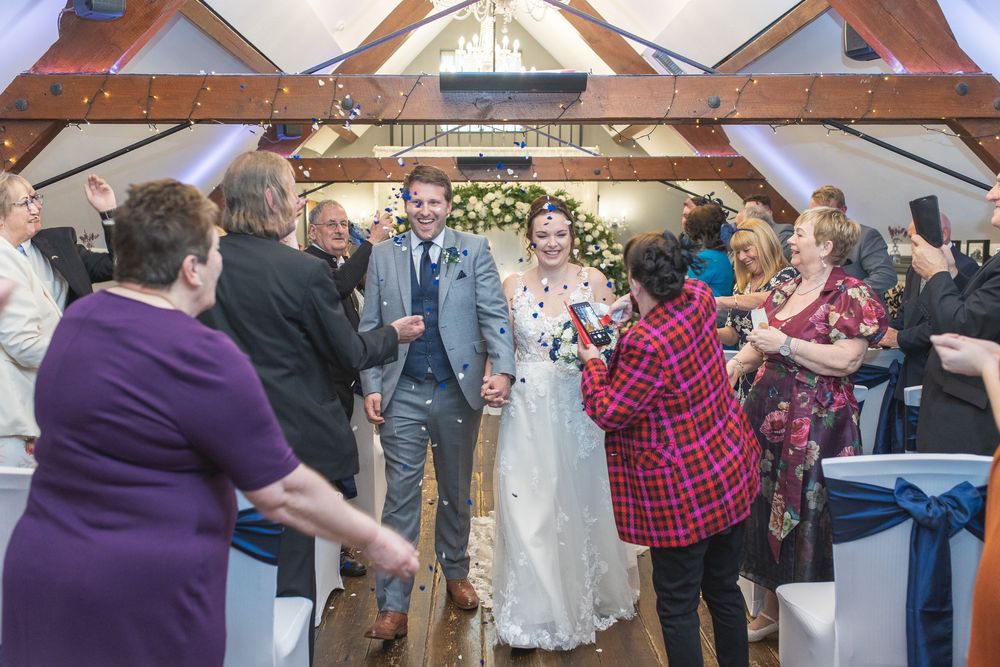 Wedding guests celebrate as newlyweds walk through a decorated indoor venue with exposed wooden beams.