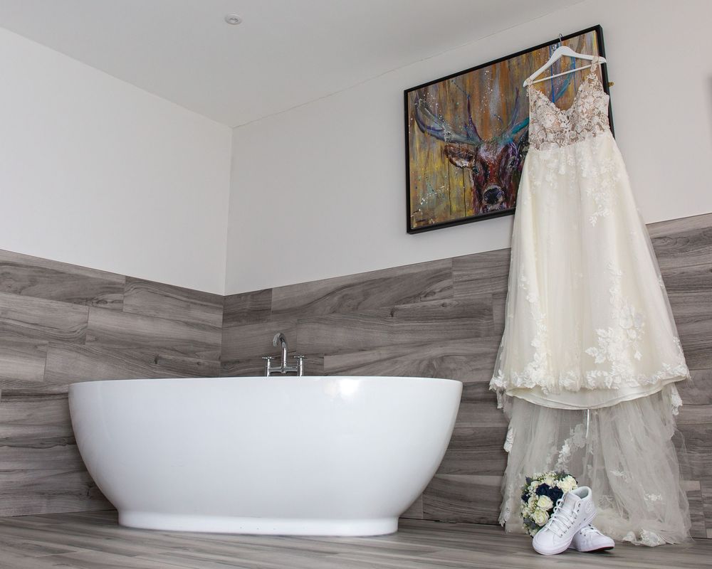 A white freestanding bathtub and lace dress are displayed in a modern bathroom with gray marble-look tile flooring.