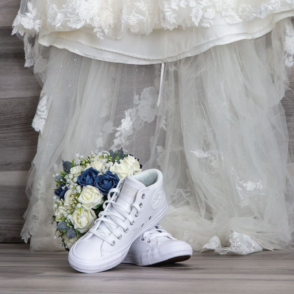 White wedding sneakers and a blue and white bridal bouquet arranged with lace wedding dress details on gray floor.