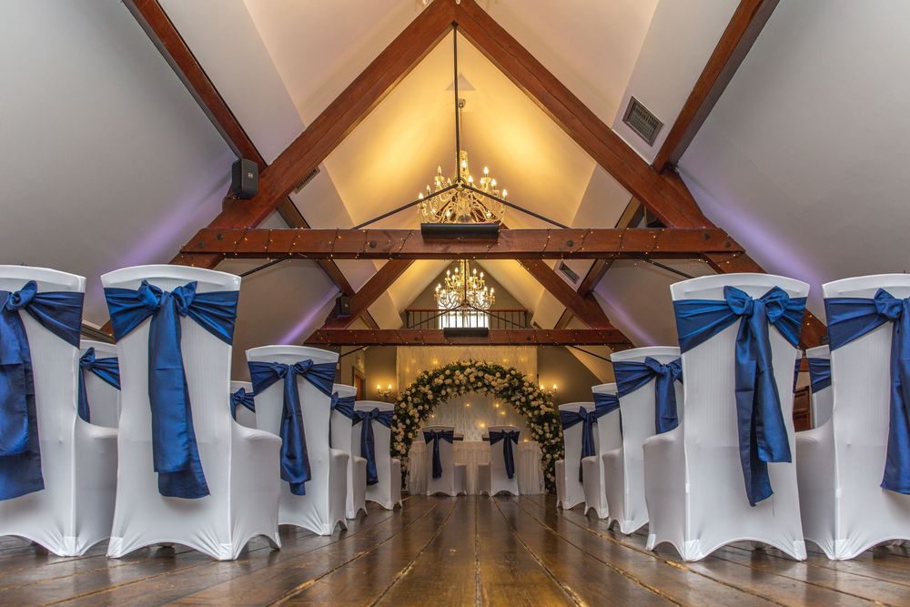 Wedding reception venue with exposed wooden beams and white chair covers decorated with blue sashes.