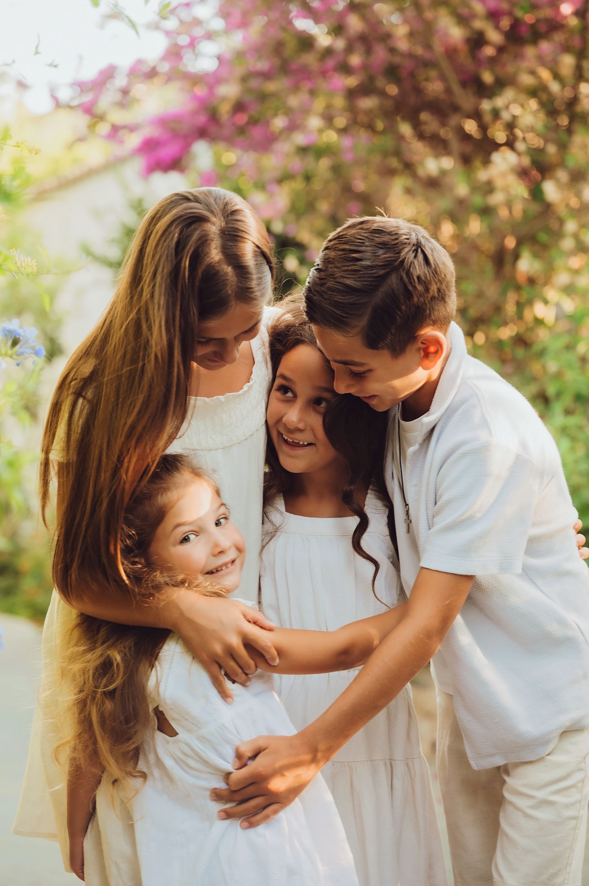 A tender family moment captured outdoors with everyone wearing white against a backdrop of pink flowering bushes.