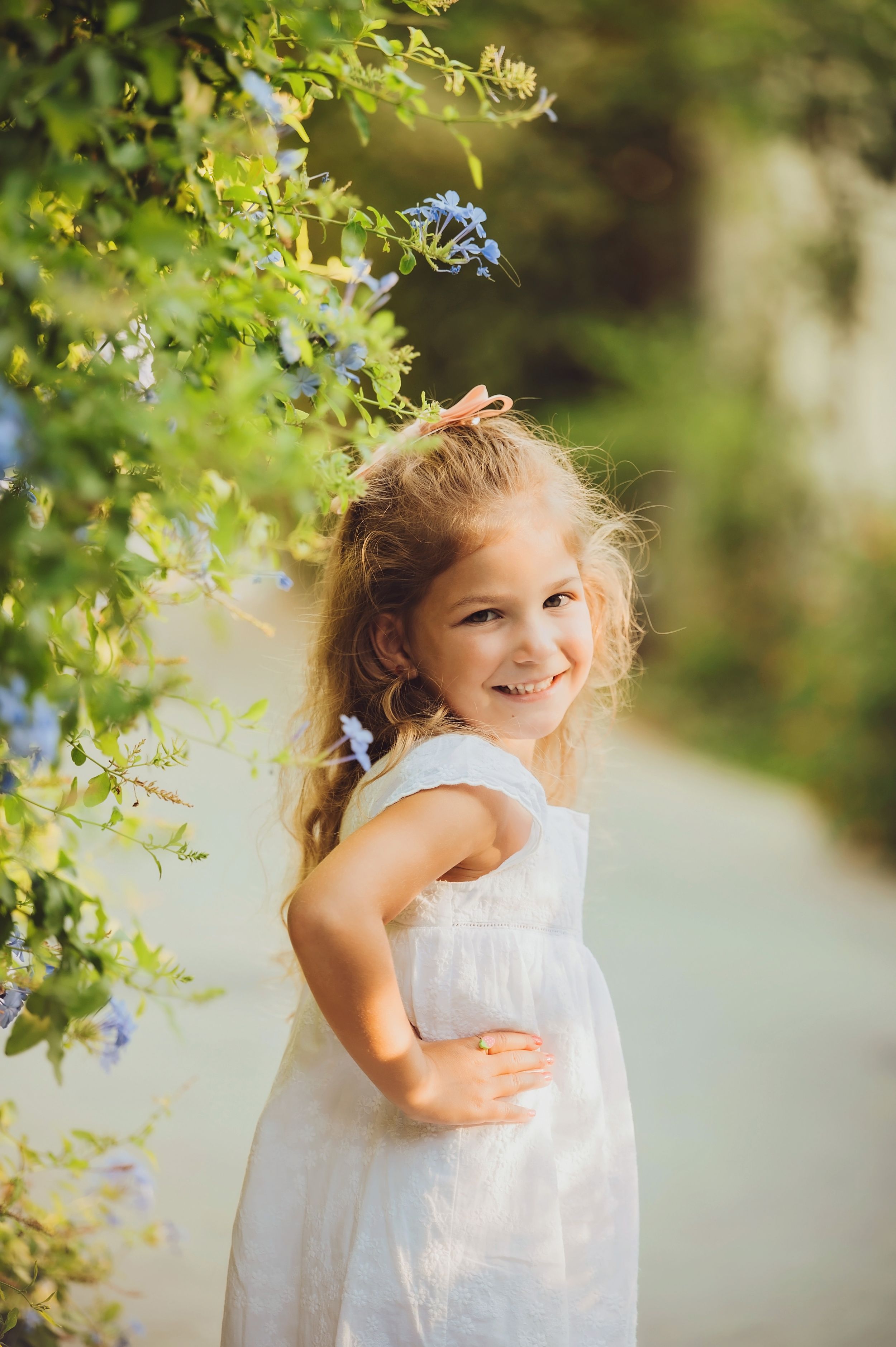 Young child in a white dress playing joyfully among green foliage in natural outdoor lighting.
